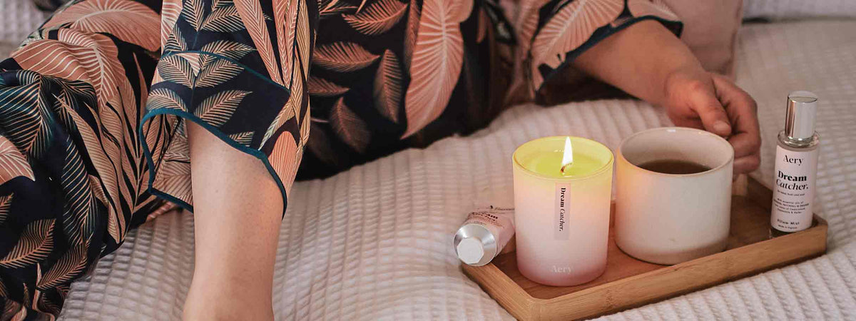 person in pyjammas relaxing in bedroom setting with scented candle, pillow spray and cup of tea
