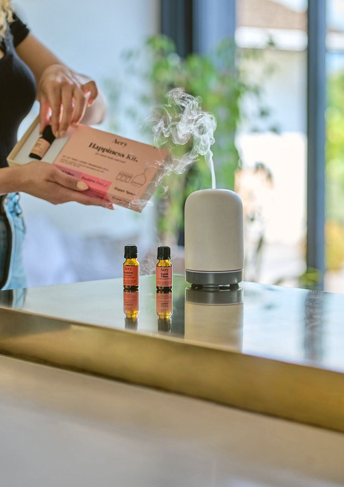 Pink Happiness set of three fragrance oils by Aery displayed with product packaging and electric diffuser placed on gold kitchen worktop